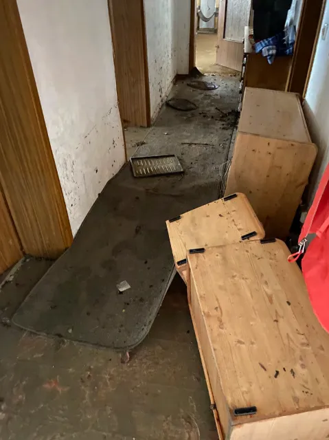 Überschwemmter Flur mit verschmutztem Teppich, umgekippten Holzmöbeln und Schlammspuren an den Wänden